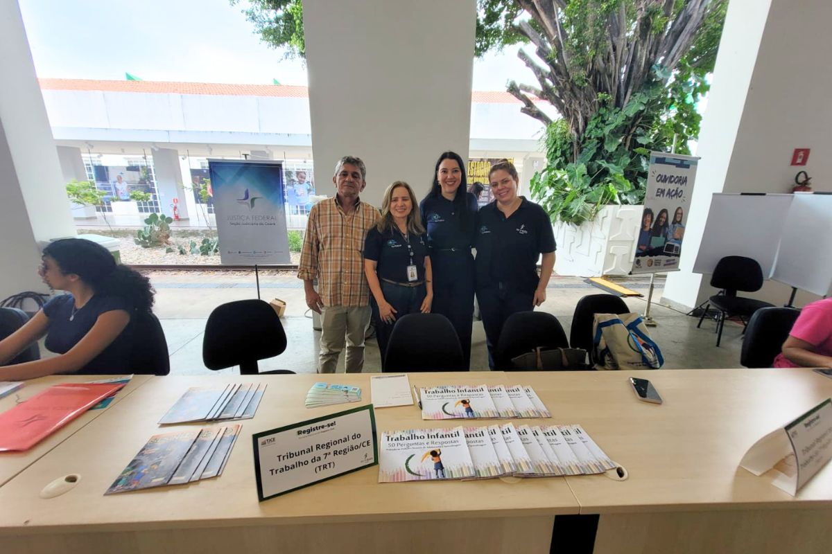 Uma fotografia em plano médio, tirada em uma área externa coberta, mostra quatro pessoas em pé atrás de uma mesa, com outros indivíduos sentados em primeiro plano e ao fundo. O grupo em pé, com três mulheres em camisas polo escuras e um homem em uma camisa xadrez, sorri para a câmera. Na mesa, há cartilhas e um pequeno banner que diz "REGISTRE-SE! Tribunal Regional do Trabalho da 7ª Região (TRT-CE)".